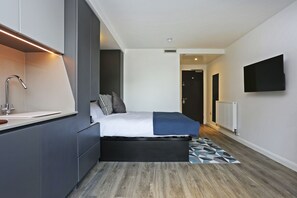 Un dormitorio moderno con kitchenette integrado, una cama con manta azul y una alfombra de estampado geométrico.