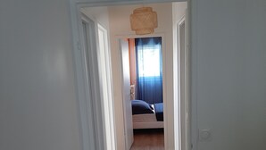 Un couloir mène à une chambre qui contient un lit, une table de chevet et une fenêtre avec des rideaux.