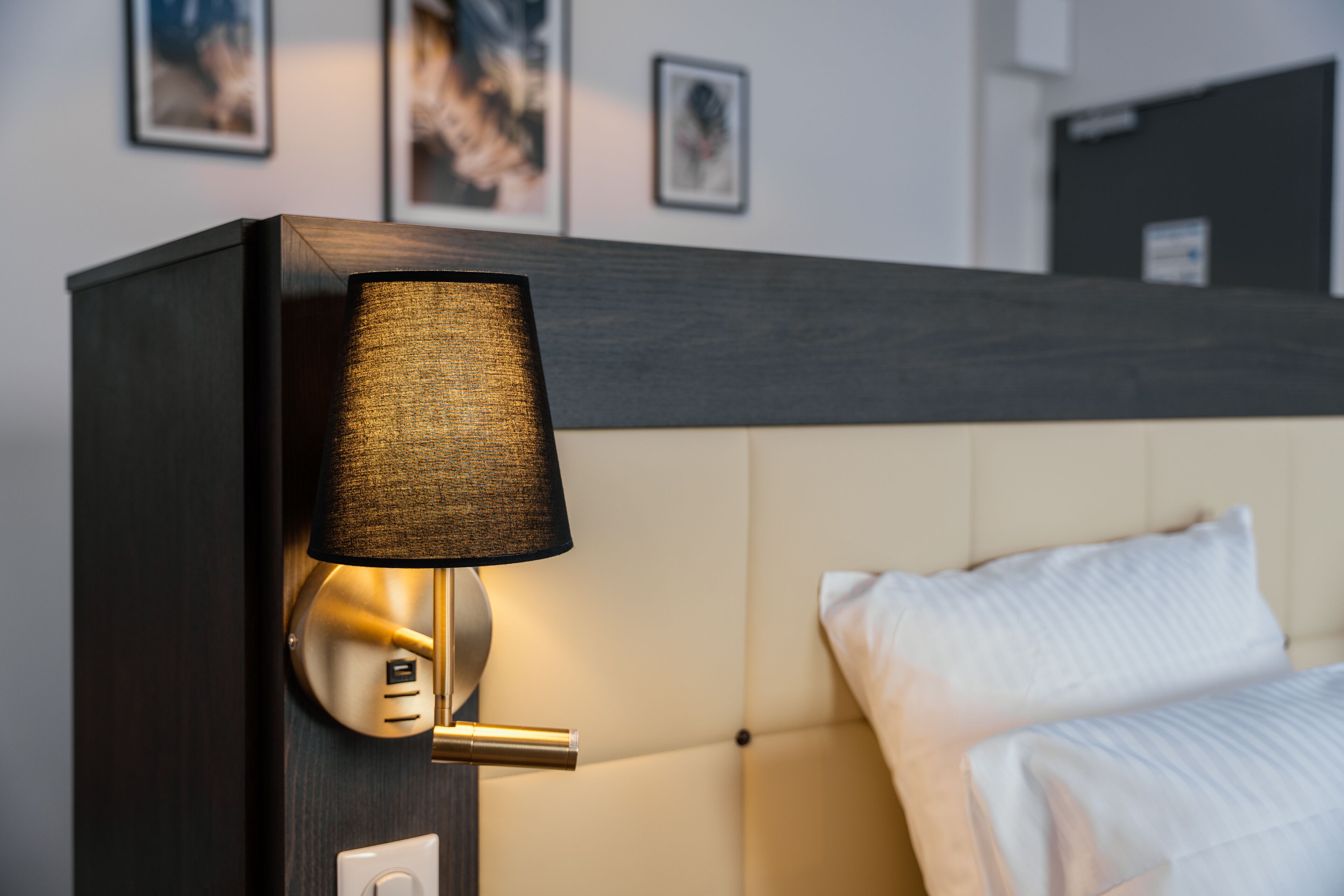 A modern hotel room with a dark wood headboard, a bedside lamp, a white bed with a textured cover, and framed artwork on the wall.