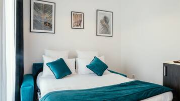 A neatly made bed with teal bedding and pillows, a dark blue headboard, and two framed artworks on the wall.