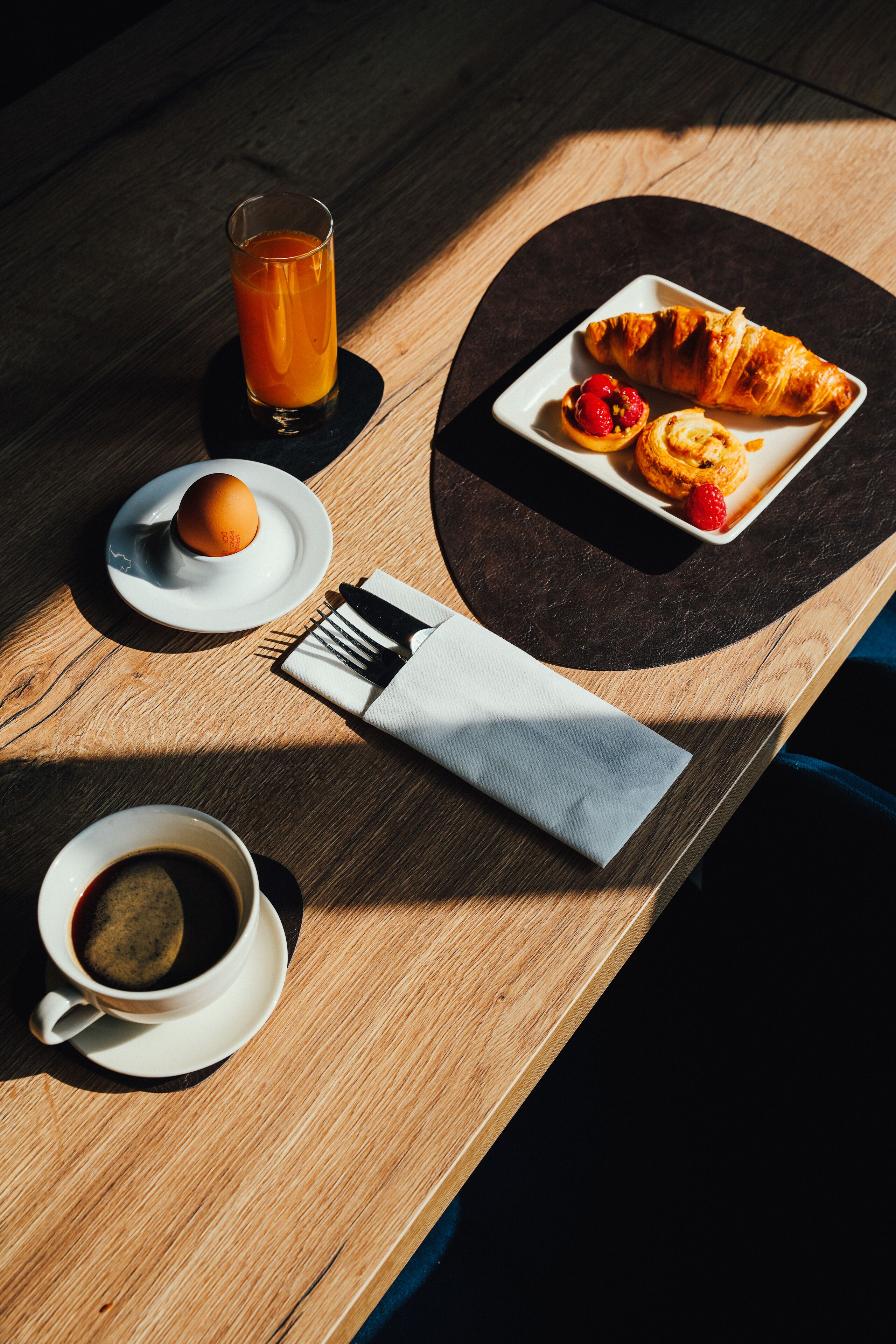 A breakfast setting with a croissant, a small plate with a berry, a glass of orange juice, and a cup of coffee on a wooden table.