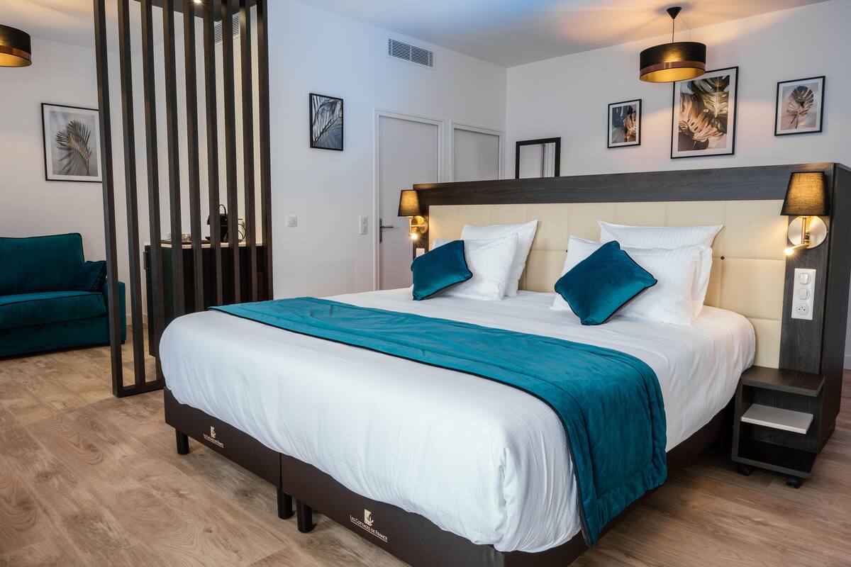 A modern hotel room with a large bed, teal bedding, a dark headboard, and wall-mounted lamps.