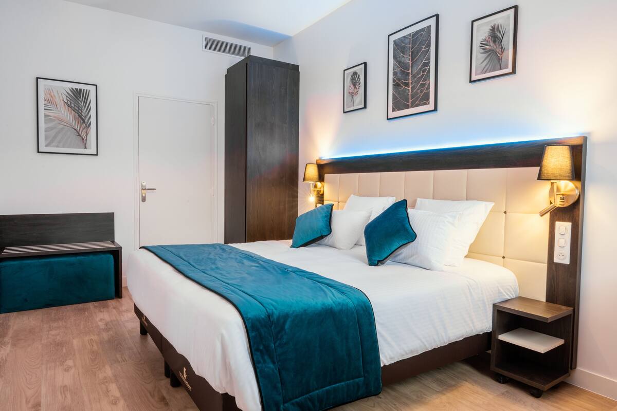 A modern hotel room with a large bed, a nightstand, a wardrobe, and framed artwork on the walls.