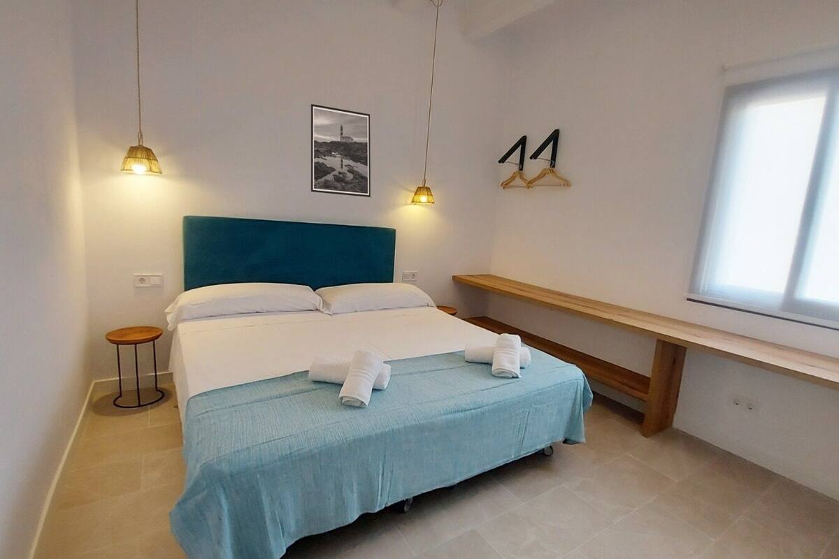 A neatly made bed with a blue bedspread and white pillows, a small round table, a bench, and a window with blinds.