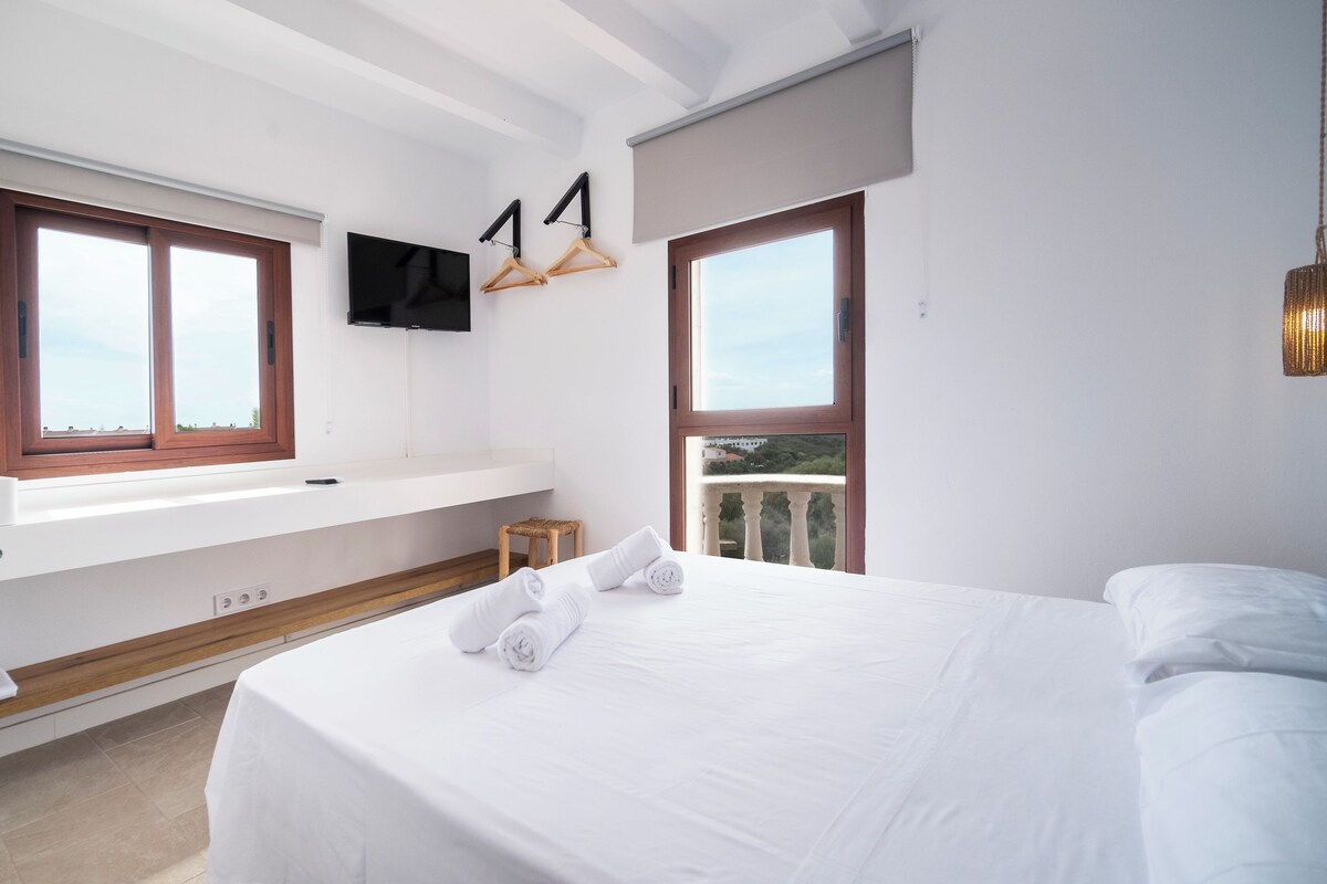 A hotel room with a large bed, a bathroom with a sink and mirror, and two windows with wooden frames.