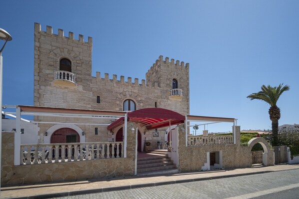 A historic stone building with a red awning, a balcony, and a palm tree nearby. - Hostal Castell de Lô (Alayor)
