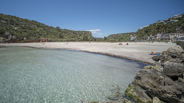 A beach with clear water, rocky shoreline, and buildings in the background. - Hostal Castell de Lô (Alayor)