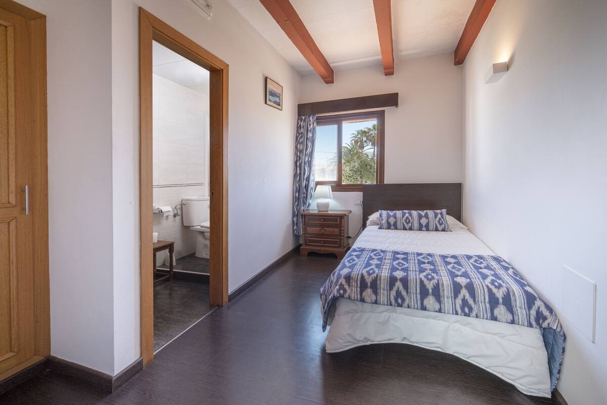 A bedroom with a bed, wooden beams, a window with curtains, and a bathroom visible through an open door.