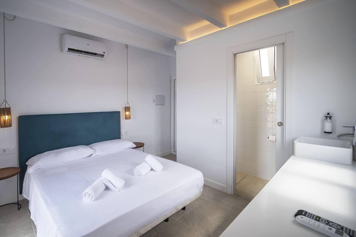 A modern hotel room with a bed, bedside tables, a bathroom with a sink and mirror, and an air conditioning unit.