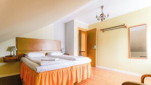 A bedroom with a wooden headboard, a bed with white linens, an orange skirted bedspread, a wooden floor, a nightstand with a lamp, a chair, a mirror, and a chandelier. - Tiglkåk - Hostel (Rauma)