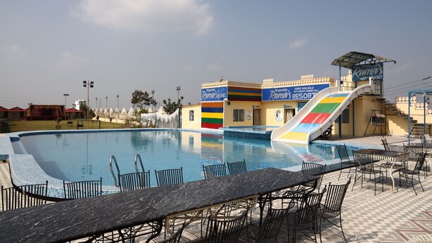 A swimming pool with a colorful slide, outdoor seating, and a building with a sign for Ram's Resort.