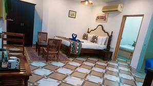 A hotel room with a bed, a desk with a chair, a small table with cups, a patterned floor, and an air conditioning unit. - Ramas Resort (Niwai)