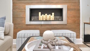 Een moderne woonkamer met een open haard, twee gestreepte poefs, een bijzettafeltje met decoratieve objecten en een bank.
