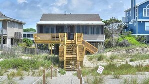 Een strandhuis met een houten trap naar een terras, omringd door zand en begroeiing.