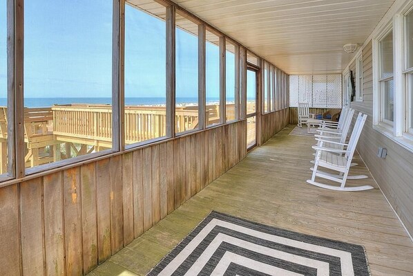 A covered porch with large windows overlooking the beach, wooden flooring, and white rocking chairs.