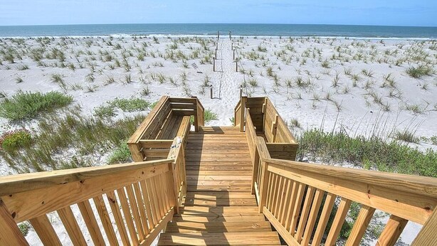 Een houten looppad dat naar een strand met zand en schaarse begroeiing leidt.
