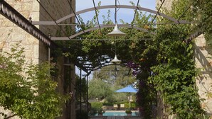 Um jardim com um caminho de madeira que leva a uma piscina, cercada por muros de pedra e vegetação.