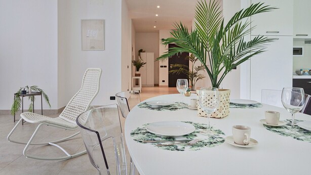 Un espace repas moderne avec une table ronde dressée pour quatre personnes, des chaises transparentes, une plante en pot et un tableau encadré au mur.