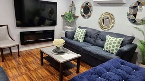 Una sala de estar moderna con un televisor de pantalla plana, un sofá de cuero con cojines verde lunares, un otomano tapizado azul, una mesa de centro de madera con un jarrón de flores y espejos decorativos en las paredes.