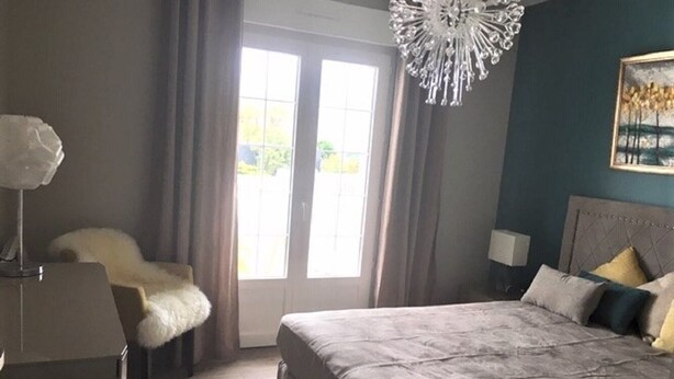 Une chambre à coucher avec un grand lit, un fauteuil recouvert d’une peau de bête, un lustre et un tableau accroché au mur.