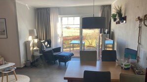 A modern living room with a dining area, a large window with curtains, a sofa, a lamp, and a potted plant.