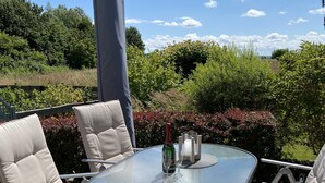A round glass table with four beige cushioned chairs, a bottle of champagne, and candles on a patio with a view of greenery and a clear sky.