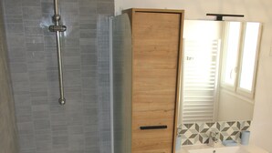 A modern bathroom with a freestanding bathtub, a wooden vanity with a mirror, and a tiled shower wall. - Charming apartment ideally located (Bayeux)