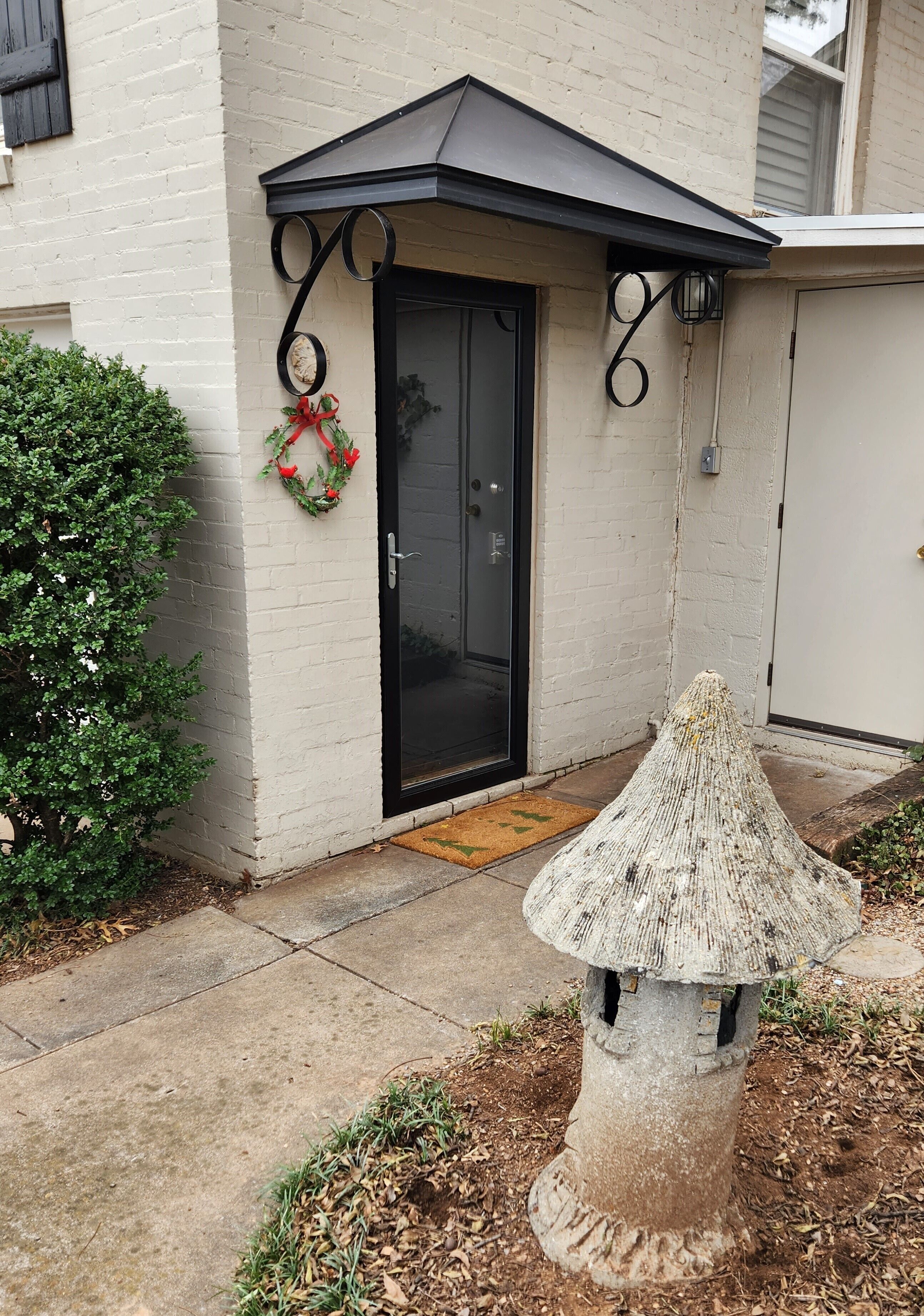 A white brick building with a black door, a small garden with a decorative stone structure, and a wreath on the door.