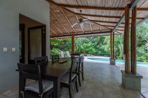 An outdoor dining area with a wooden table and chairs, a ceiling fan, and a view of a swimming pool and lush greenery. - Casa Escondida | Serene Retreat with Pool (Nosara)