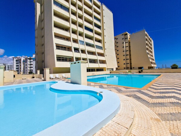 A swimming pool area with a curved pool, lounge chairs, and a high-rise building in the background. - Apartment Praia Rocha | Sea & Pool | Sunny (Portimão)