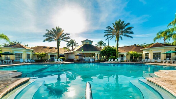 A large swimming pool with lounge chairs and palm trees, surrounded by buildings with a Mediterranean architectural style.