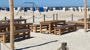 Ein Strandbereich mit Holztischen, Bänken, einem Seilabfangnetz und einer Schaukel.