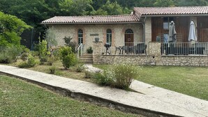 Un edificio de piedra con techo de tejas, un patio con mobiliario exterior y una zona ajardinada.