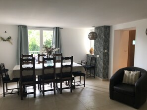 A dining area with a table set for six, surrounded by wooden chairs. A vase with flowers is on the table, and a hanging light fixture is above.