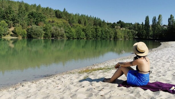 Una persona sentada en una playa arenosa cerca de un lago tranquilo, con árboles y un cielo despejado al fondo.