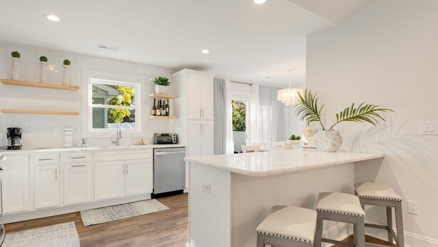 Una cocina moderna con armarios blancos, una isla central con taburetes y estanterías empotradas con plantas y botellas.