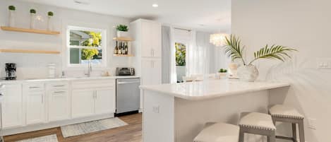 A modern kitchen with white cabinetry, a central island with bar stools, and built-in shelves with plants and bottles.