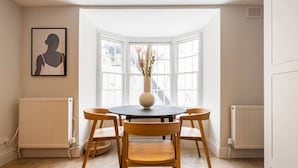 Een eetruimte met een ronde tafel, vier houten stoelen, een vaas met bloemen en een kunstwerk in een lijst aan de muur.