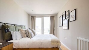 A bedroom with a bed, bedside table, curtains, and framed pictures on the wall. - The Nine Elms Lane Arms - Stunning & Bright 2bdr Flat With Balconies (London)