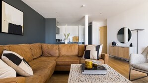 A modern living room with a brown leather sofa, a wooden coffee table, and a grey armchair. A kitchen is visible in the background. - The Nine Elms Lane Arms - Stunning & Bright 2bdr Flat With Balconies (London)