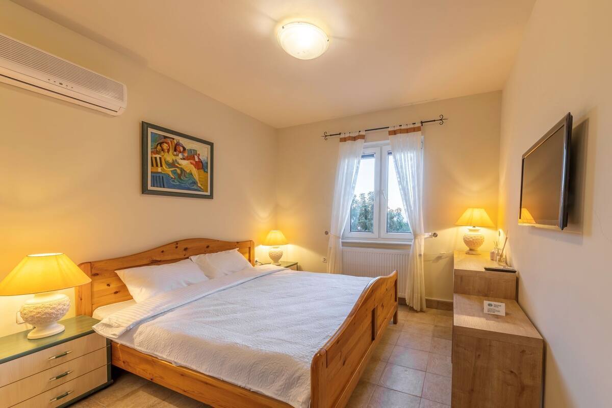 Een slaapkamer met een houten bed, twee bedlampen, een schilderij aan de muur, een raam met gordijnen en een aan de muur bevestigde televisie.
