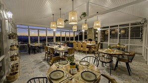 A dining area with round tables, wicker pendant lights, and a view of the city at sunset. - Apa Mountain Lodge (Konyaalti)