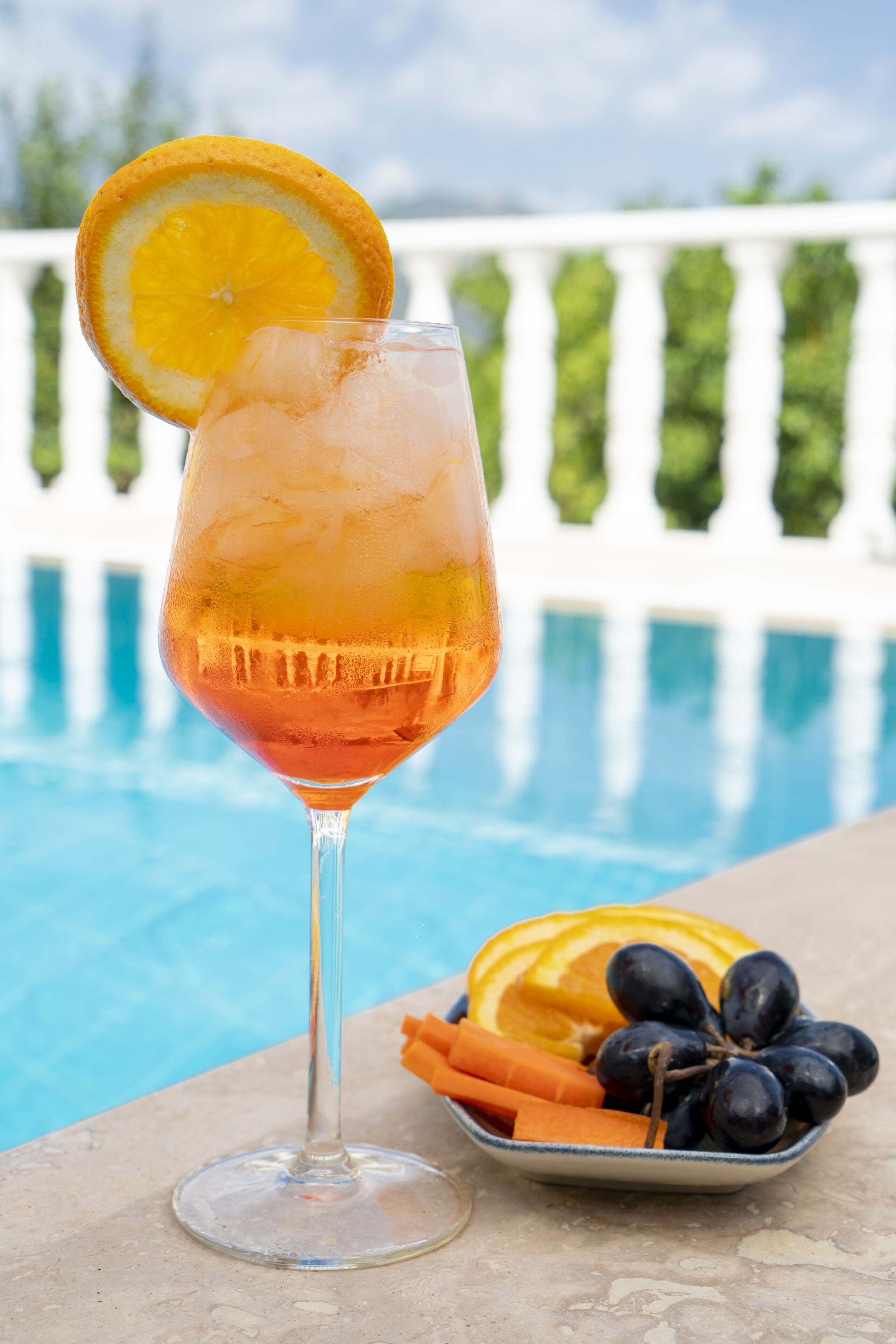 A cocktail glass with a slice of orange on the rim, placed on a terrace with a pool in the background.