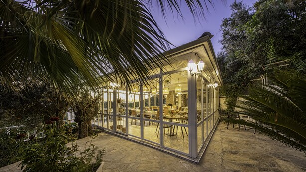 A glass-enclosed building with a stone floor, surrounded by greenery and palm trees. - Apa Mountain Lodge (Konyaalti)