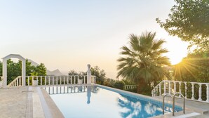 A swimming pool with a white balustrade, surrounded by greenery and palm trees, with a clear view of the sky and mountains in the background. - Apa Mountain Lodge (Konyaalti)