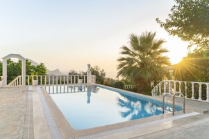 A swimming pool with a white balustrade, surrounded by greenery and palm trees, with a clear view of the sky and mountains in the background.