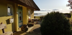 Eine Holzterrasse mit Tisch und Stühlen, ein Reetdach und Blick auf einen See.