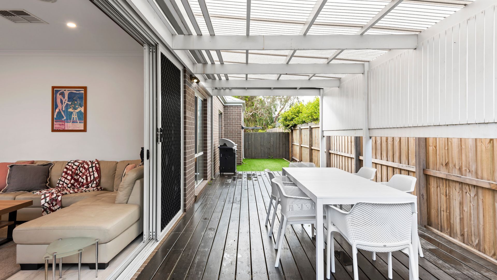 En overdækket terrasse med træterrasse, et hvidt bord og stole, en sofa og en grill.