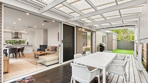 Una cocina moderna con armarios blancos, un comedor con mesa y sillas blancas, y un patio cubierto con una parrilla.