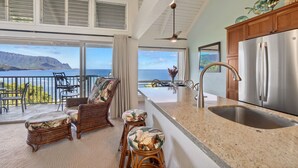 A modern kitchen with a stainless steel refrigerator, a large island with a sink, and a view of the ocean. - Pali Ke Kua #222 - Breathtaking Ocean Views! (Princeville)